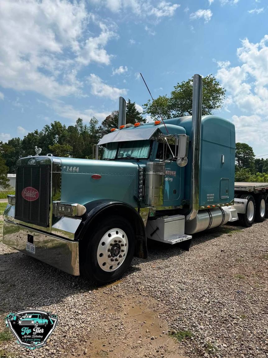A teal Peterbilt semi truck with polished chrome stacks parked on gravel under a blue sky
